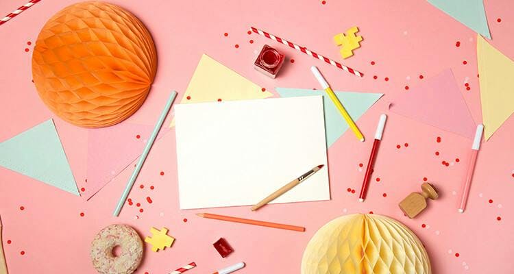 Pembe renk arka plan üzerinde; parti malzemeleri, renkli kalemler, kesilmiş renkli kağıtlar, boya ve boya fırçası bulunuyor.