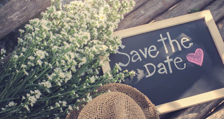 Ahşap bir arka plan üzerinde küçük boyutta siyah bir tahta, şapka ve çiçek demeti bulunuyor. Tahtanın üzerinde "Save the Date" yani "Tarihi Kaydet" yazıyor.